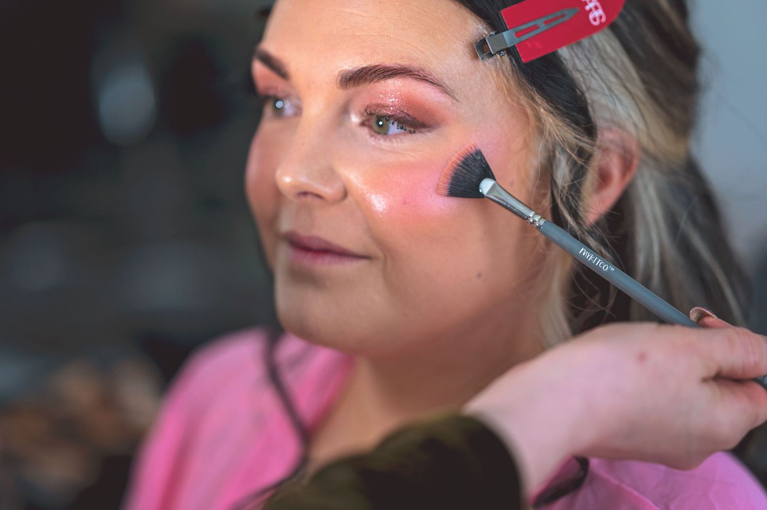 Professional makeup artist applying blush to a young blonde woman wearing a pink top in a beauty salon.