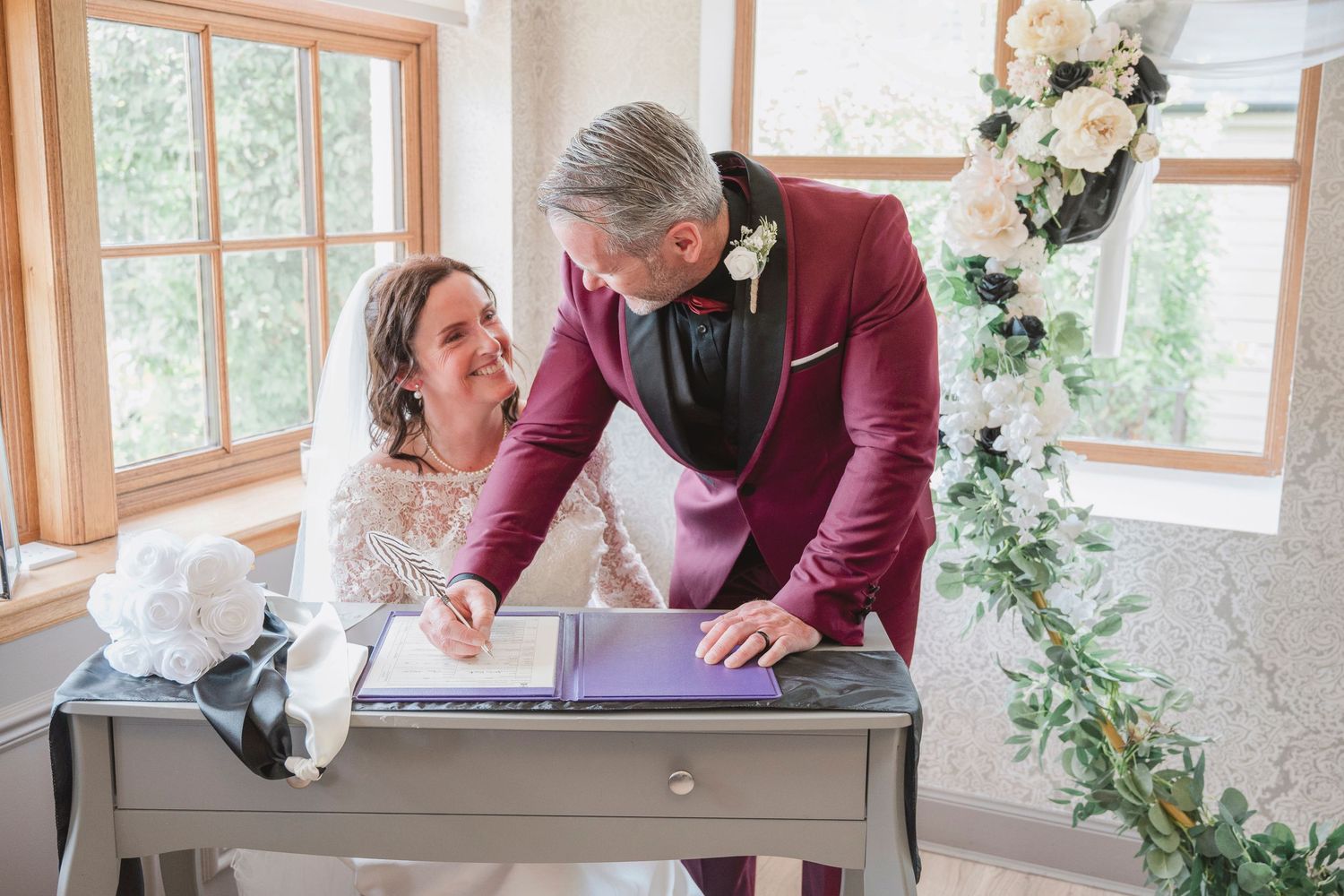 Bride and groom signing wedding register at decorated table, groom in burgundy suit, bride in white lace dress.