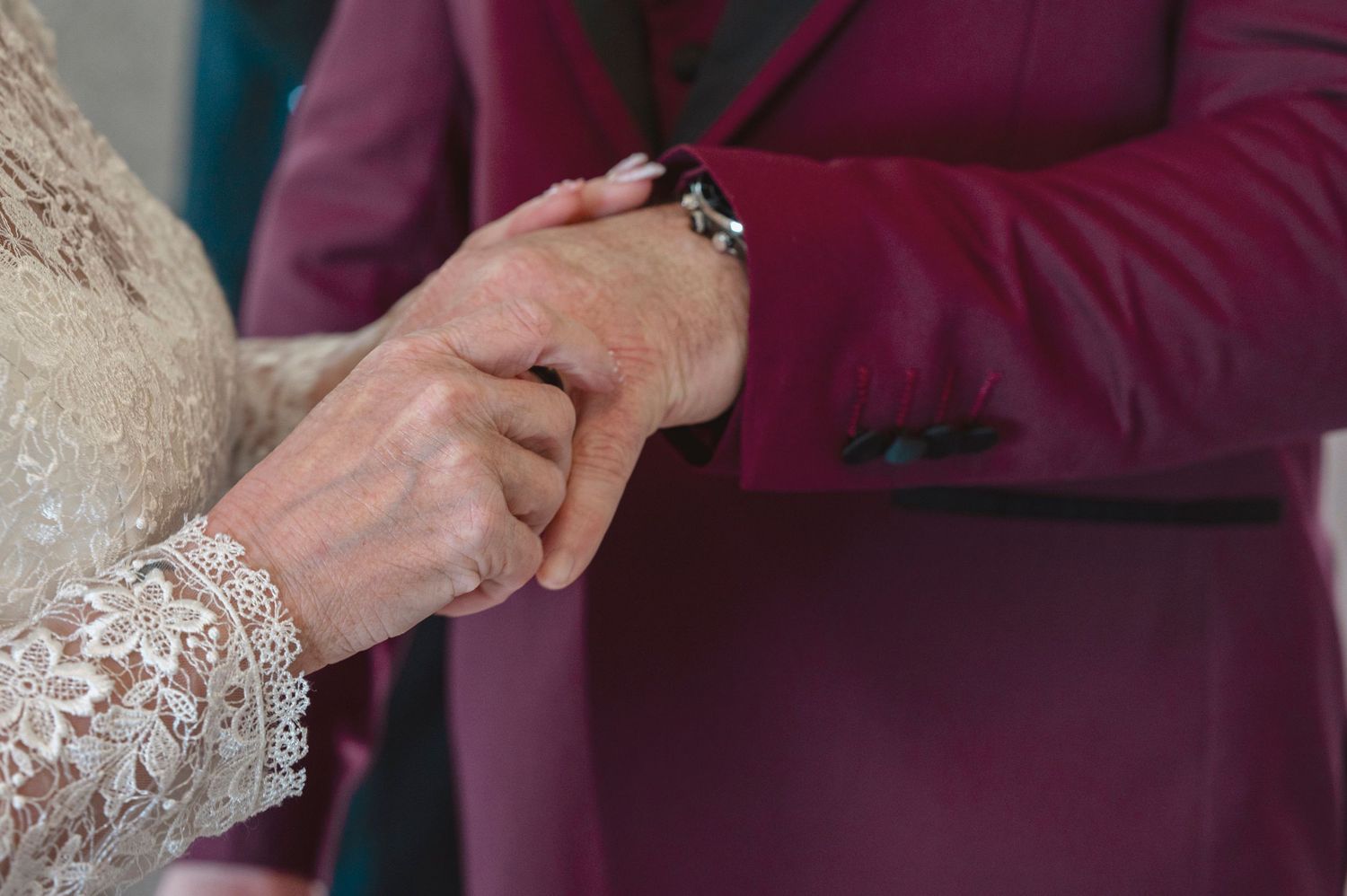 Elderly couple holding hands during wedding ceremony, bride in lace dress, groom in burgundy suit.