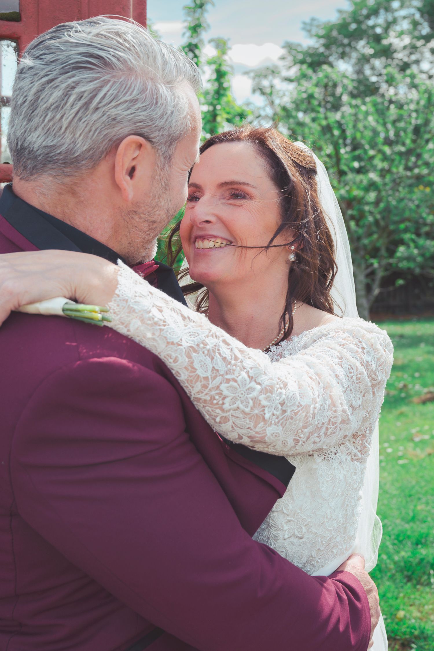 Smiling bride in lace wedding dress embraces groom in burgundy suit outdoors on their wedding day.