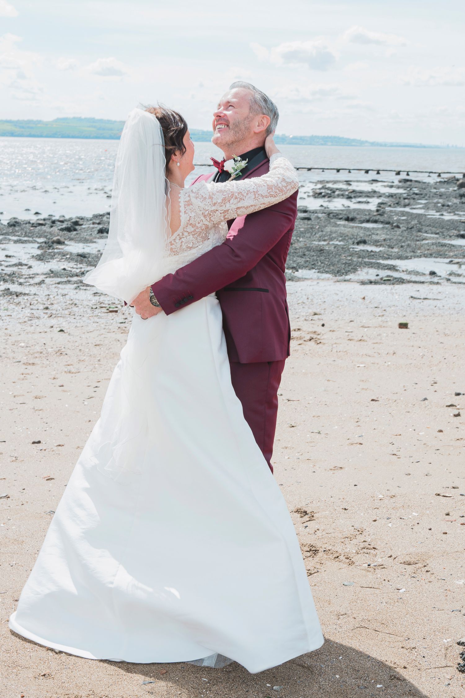 Bride and groom embracing on a sandy beach, wearing white wedding dress and burgundy suit, with ocean views behind them.