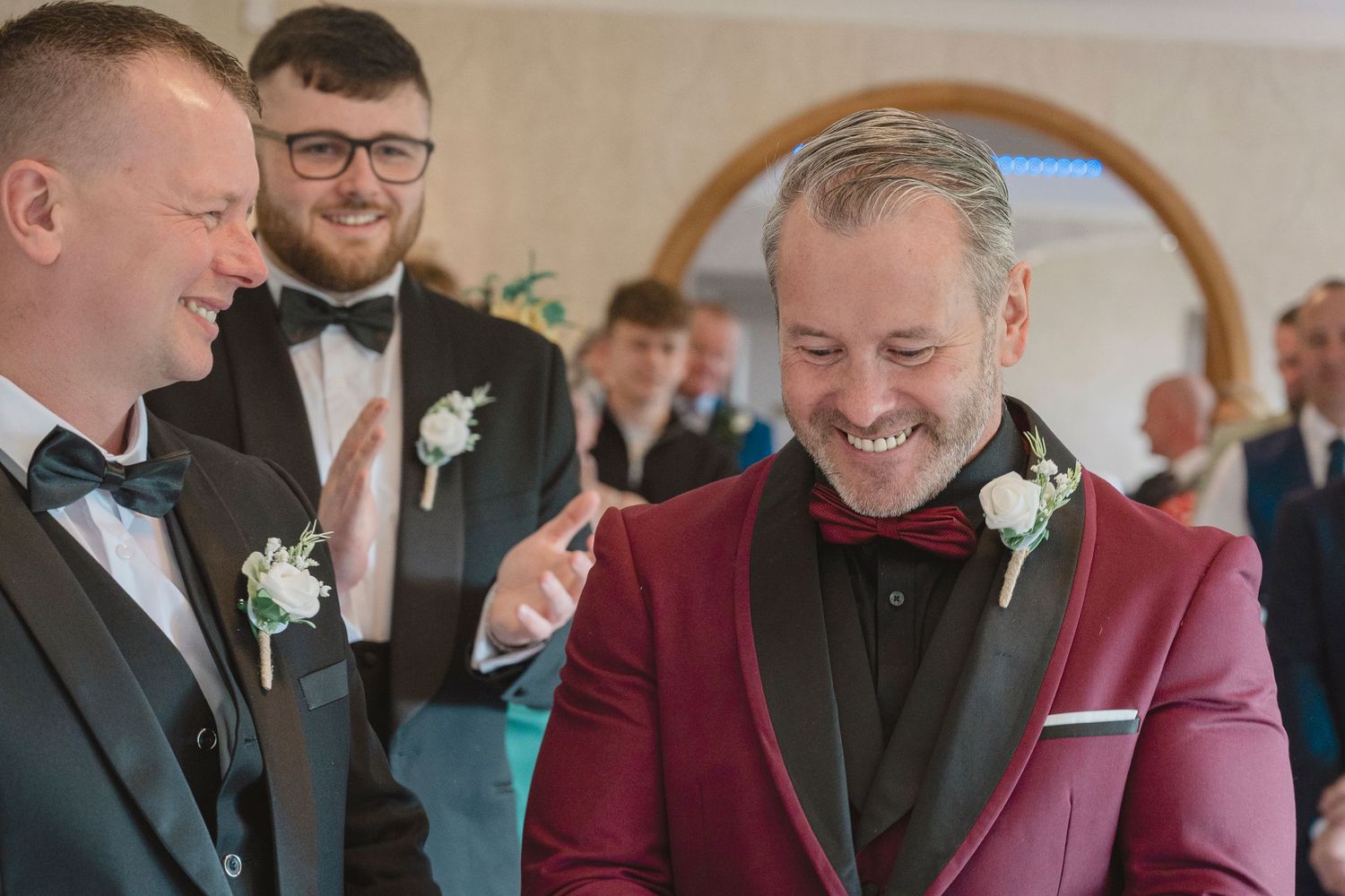 Two grooms share a joyful laugh at their wedding ceremony, wearing a burgundy tuxedo and black suit with boutonnieres.
