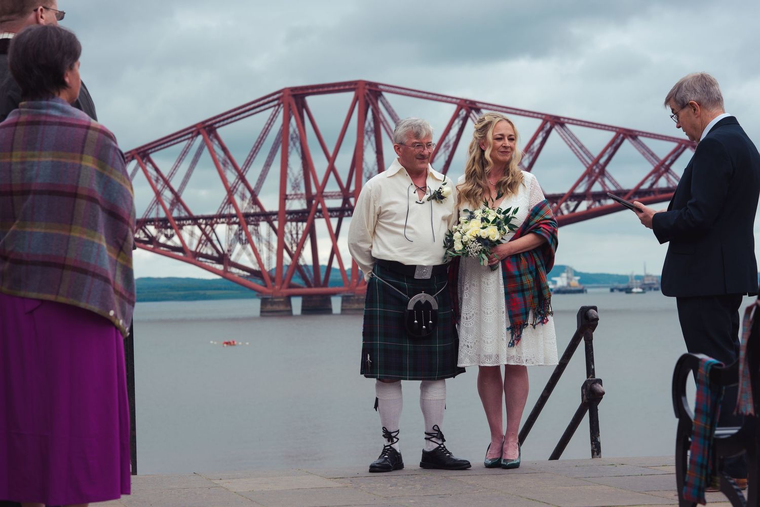 Couple poses at outdoor wedding ceremony by the Forth Bridge in Scotland, man in kilt holding bouquet.