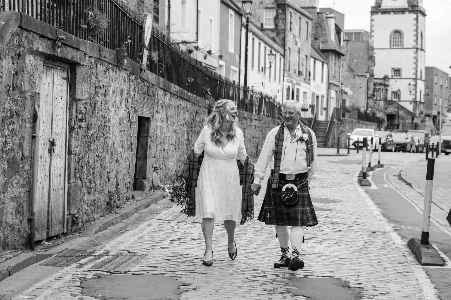 Black and white photo of a bride in white dress and groom in kilt walking hand in hand on cobblestone street.