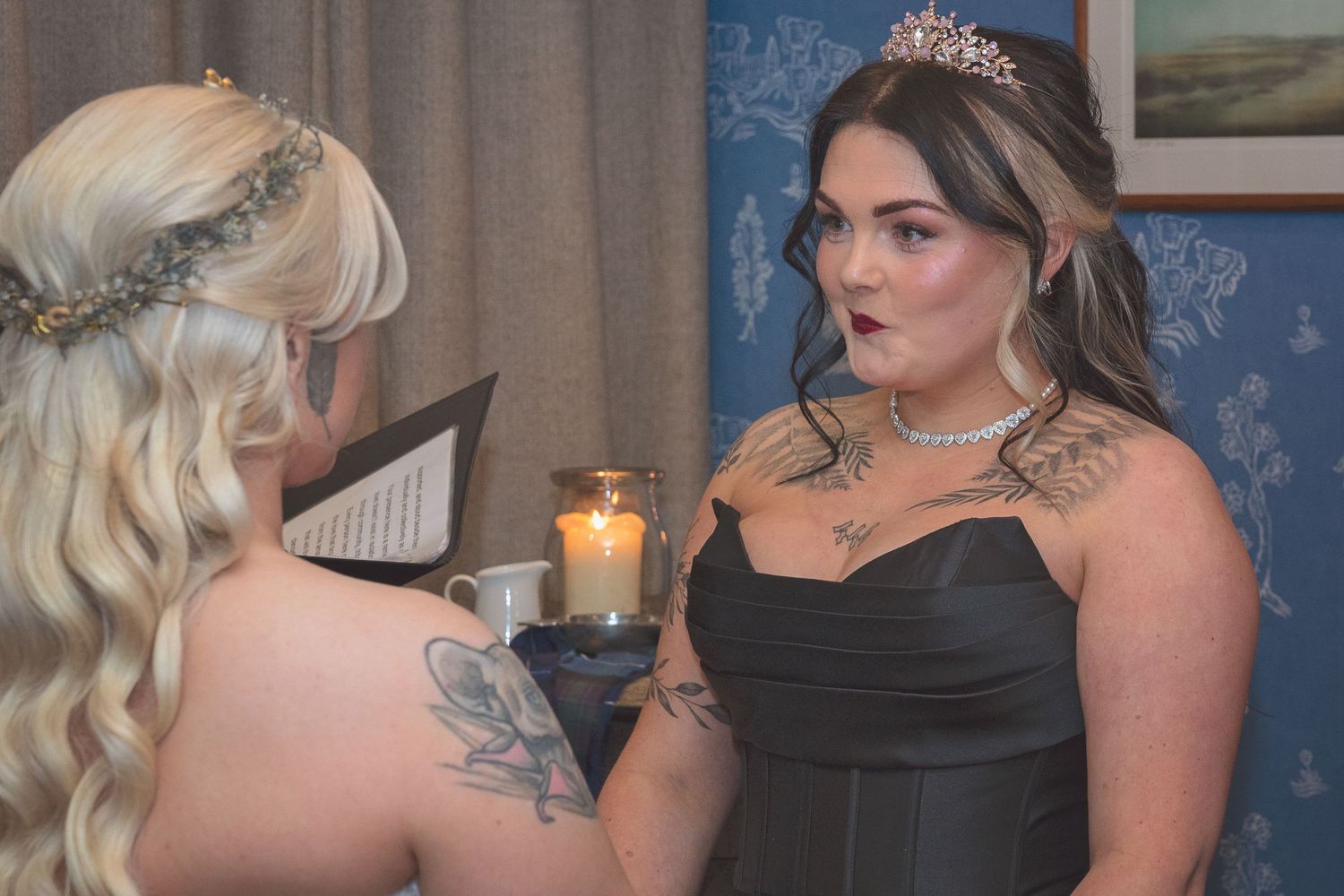 Two women in elegant black gowns with tattoos share an intimate moment by candlelight at a wedding.
