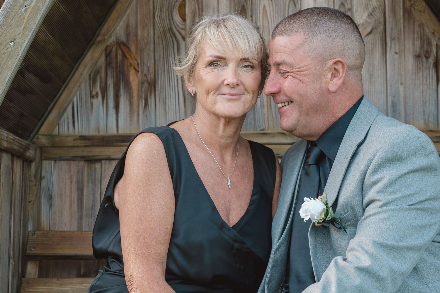 Couple smiling together in formal attire near rustic wooden wall, man wearing boutonniere, woman in elegant black dress.