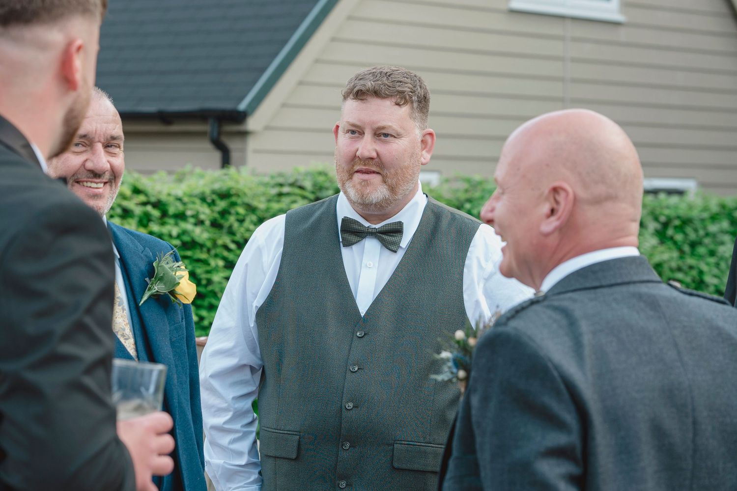 Groom in grey waistcoat and bow tie smiling with guests at an outdoor wedding celebration.