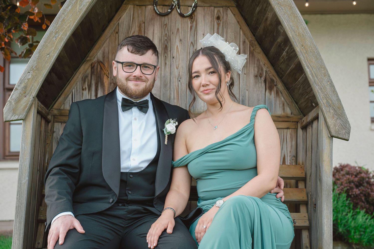 Couple in formal attire sitting in wooden arbor, man in tuxedo and woman in green gown at wedding.
