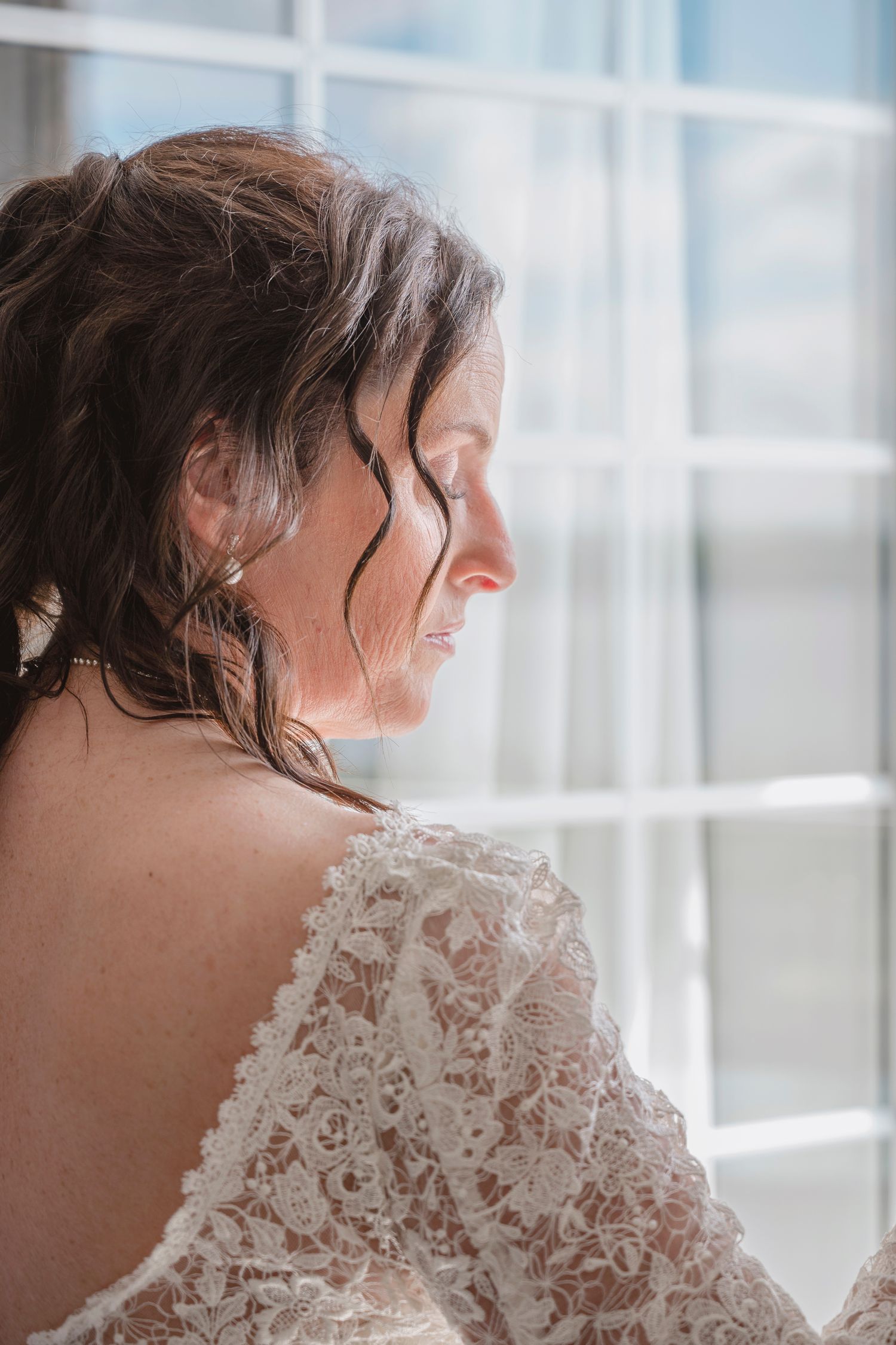 A bride in a lace wedding dress gazes downward near a bright window, showcasing her elegant updo and bare back.