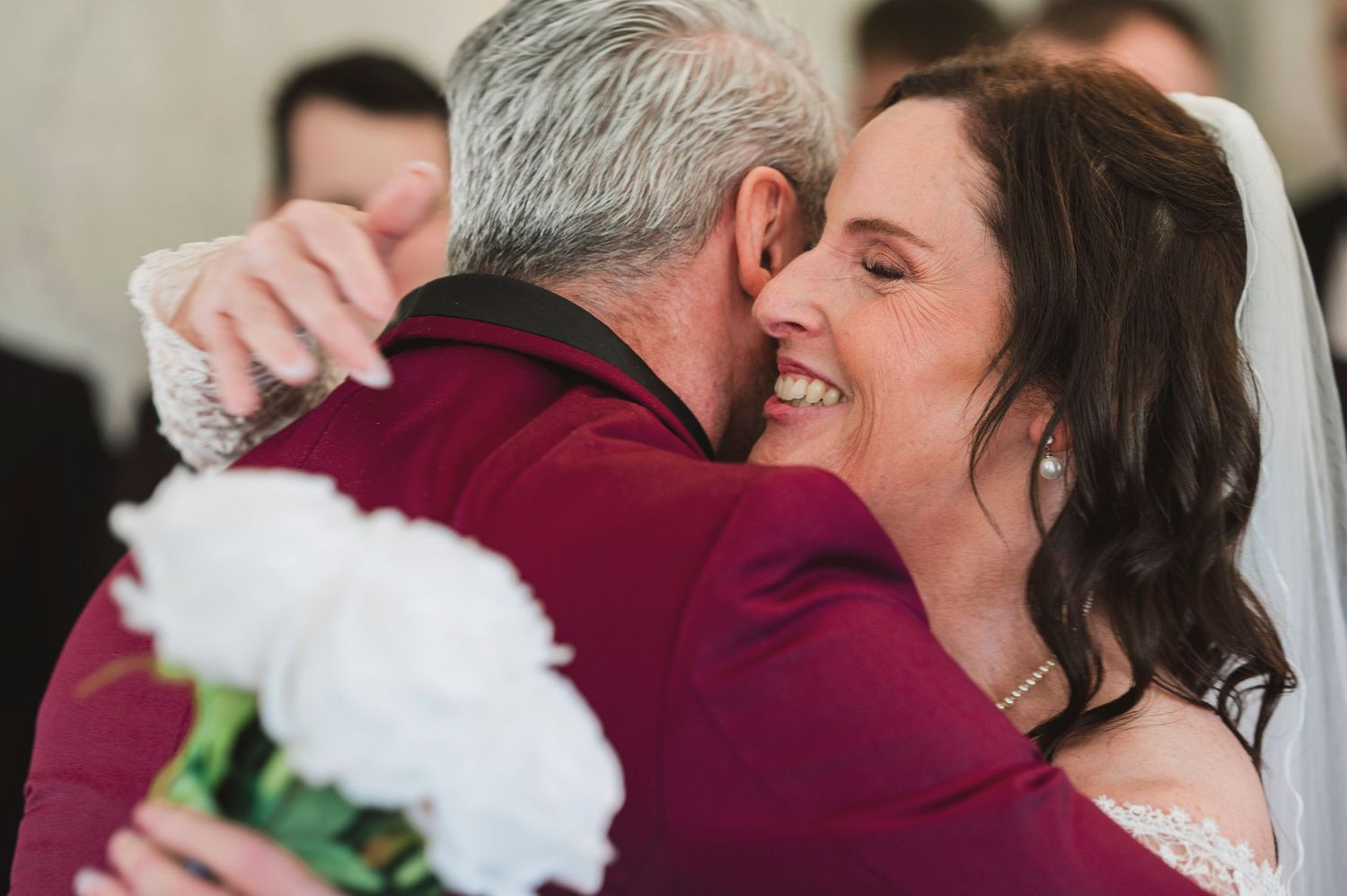 Bride and groom share a joyful embrace during wedding ceremony, bride holding white peony bouquet in elegant lace gown.