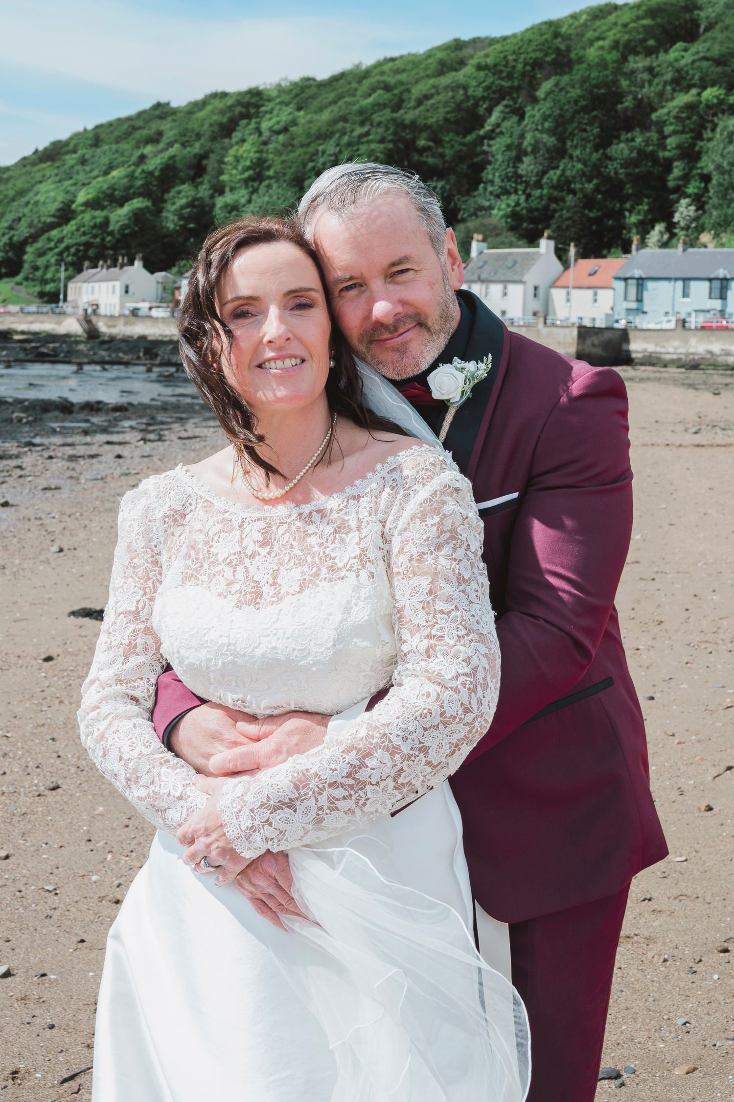 Bride and groom embrace on a sandy beach, wearing white lace gown and burgundy suit, with a coastal village backdrop.