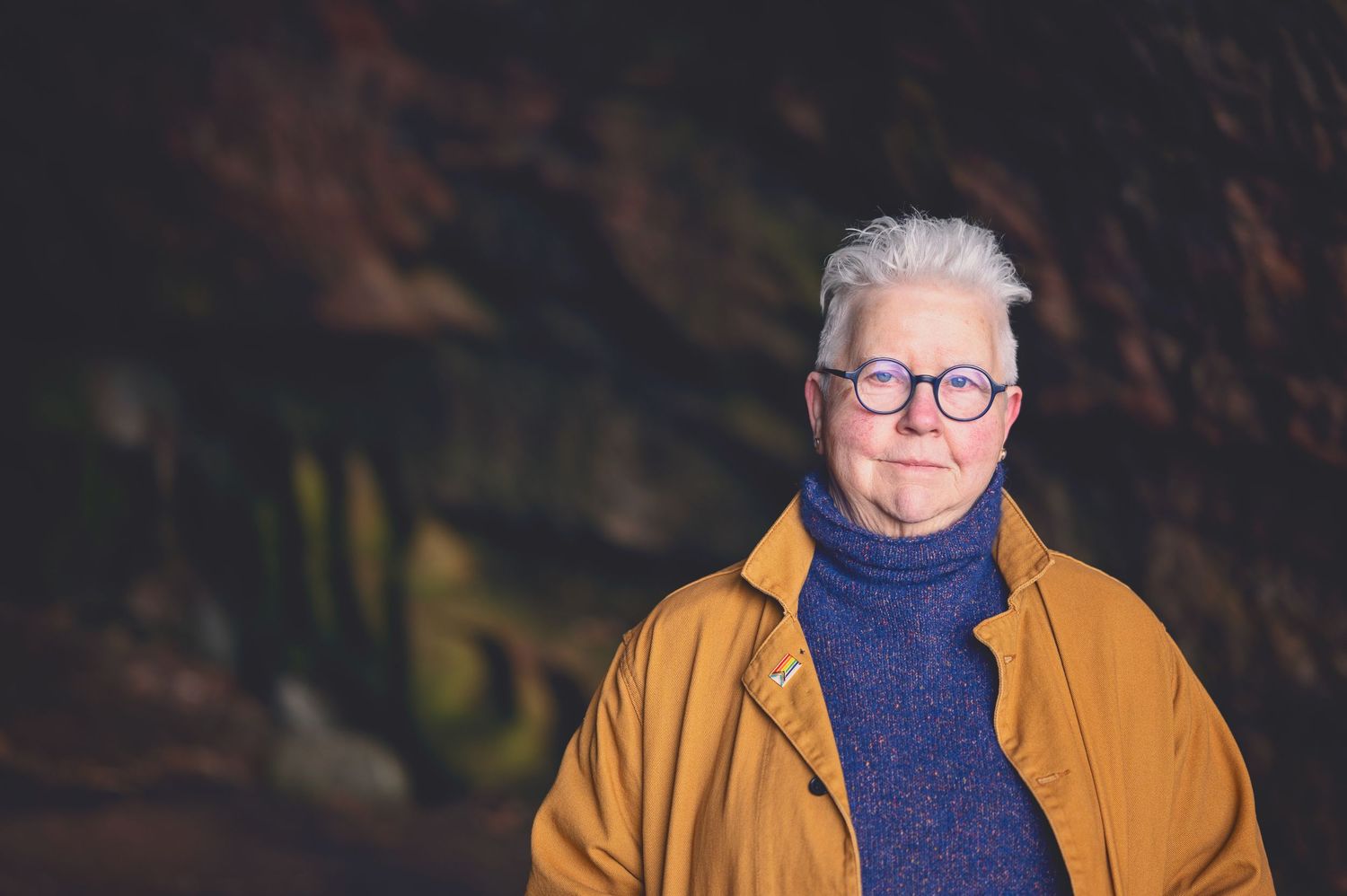 The Author Val Mcdermid with short white hair and round glasses wearing a mustard jacket and blue turtleneck sweater in the Weymss caves