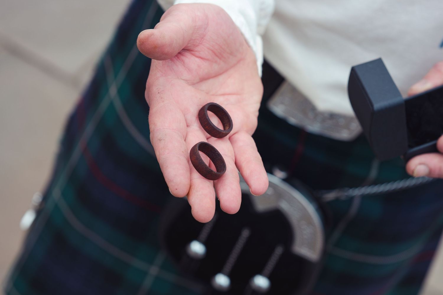 Person in tartan kilt holding two dark wooden wedding rings in palm, with ring box visible nearby.
