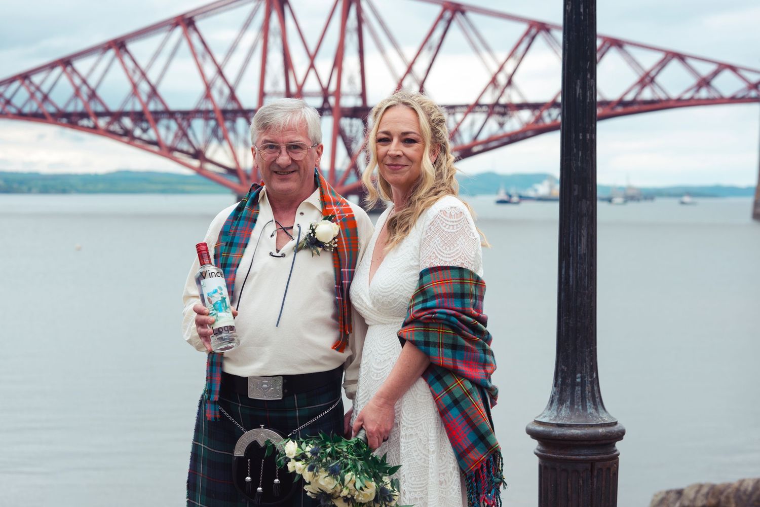 Couple posing in front of the Forth Bridge in Scotland, groom in tartan kilt, bride in white dress with plaid shawl.
