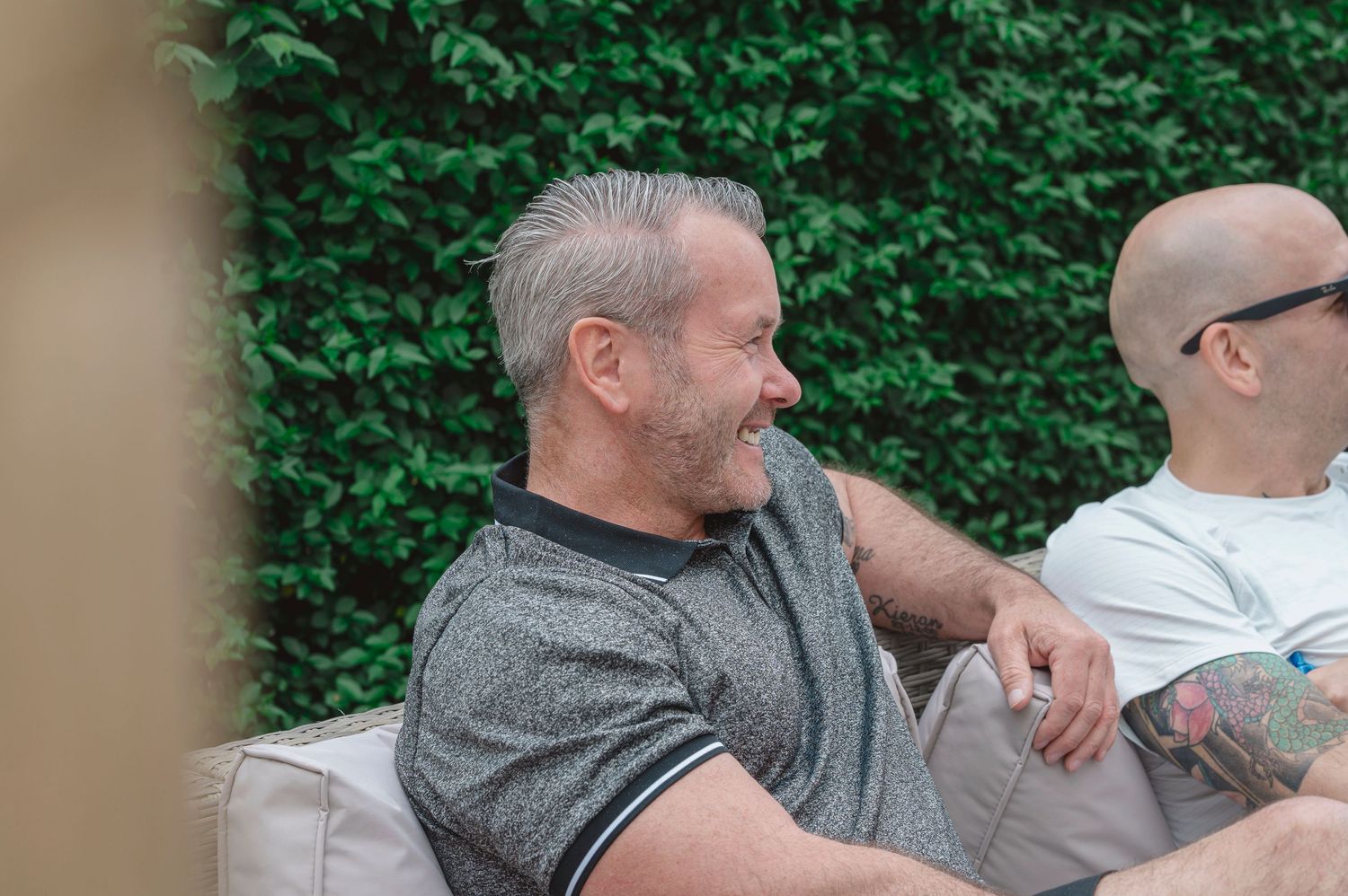 Two men sitting outdoors on a couch, smiling, with green foliage in the background.