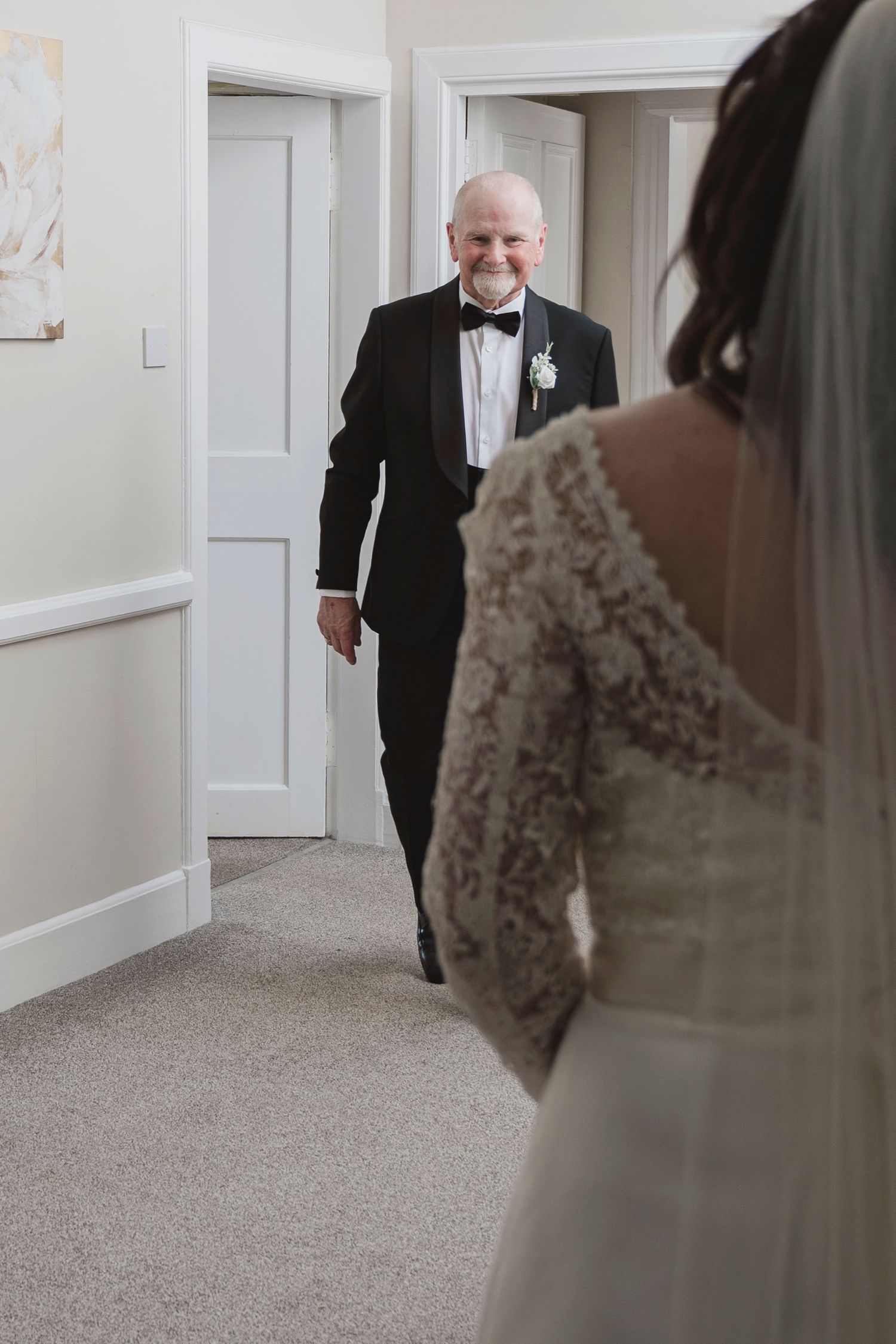Father in tuxedo smiling as he sees bride in lace wedding dress during emotional first look moment.