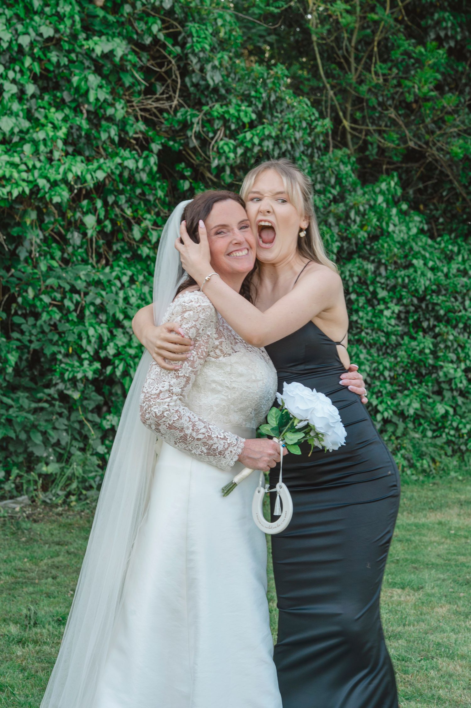 Bride in lace gown and veil hugs smiling bridesmaid in black dress holding white peony bouquet outdoors.