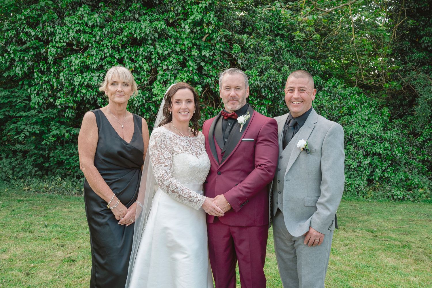 Wedding party of four posing outdoors, bride in white lace gown, groom in burgundy suit, guests in gray formal attire.