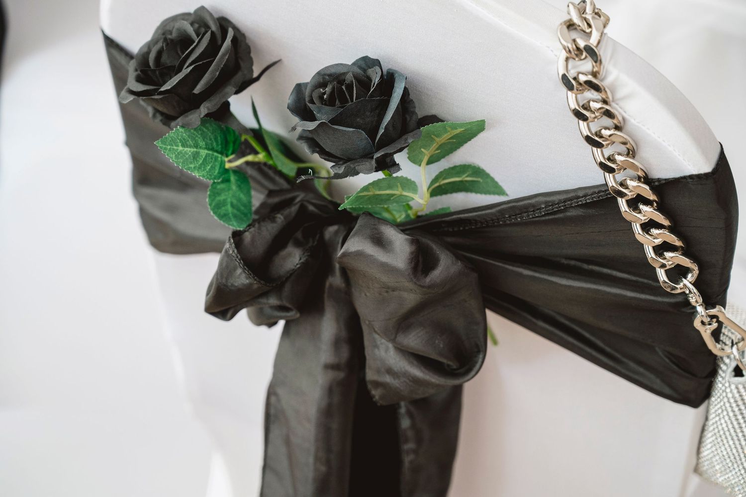 Black satin bow with artificial black roses and green leaves decorating a white chair at a formal event.