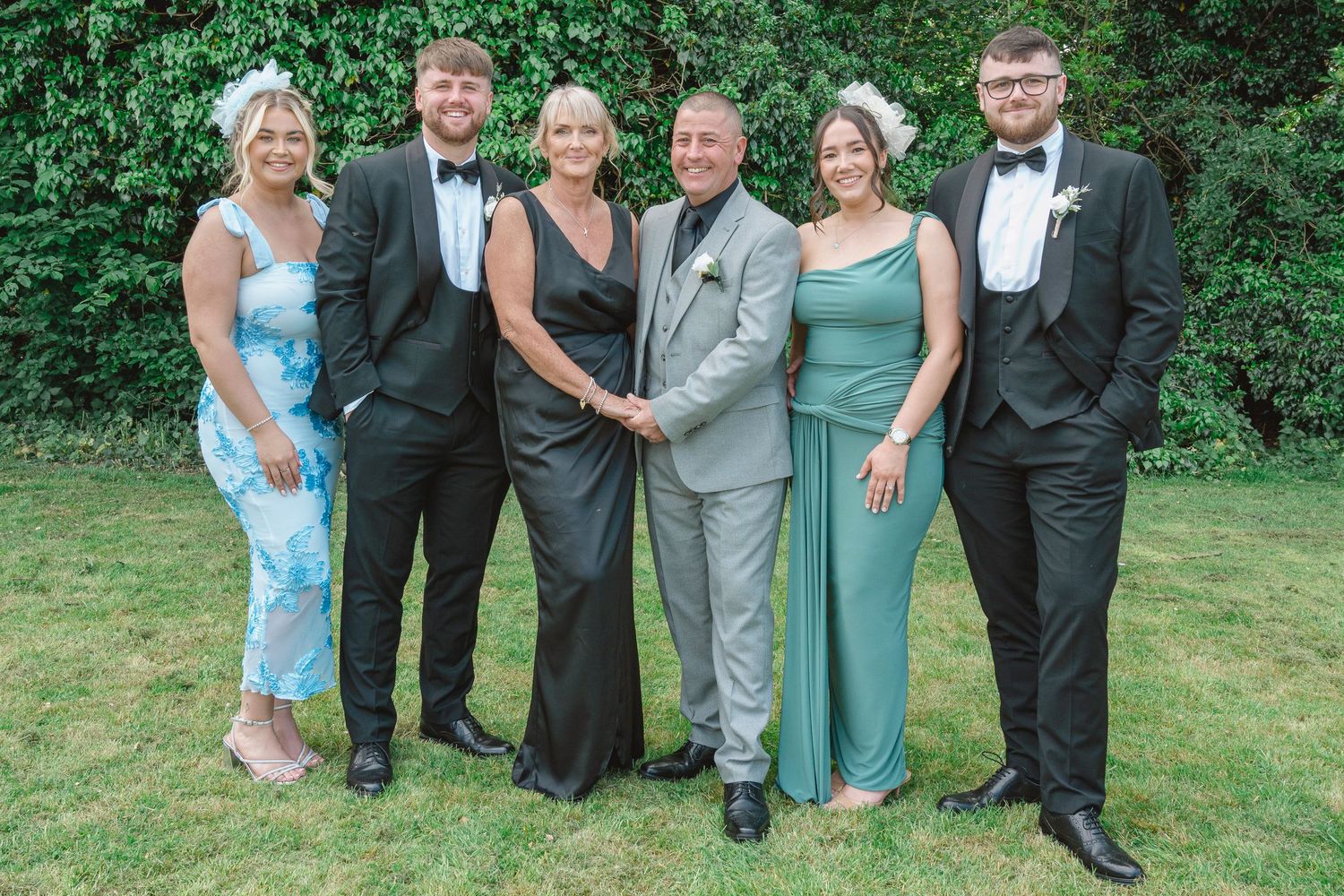Six elegantly dressed people posing together outdoors at a formal event, wearing black tie and evening gowns on green lawn.