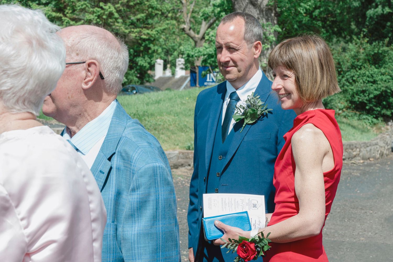 Elegantly dressed wedding guests in blue suit and red dress socializing outdoors at a sunny summer wedding ceremony.