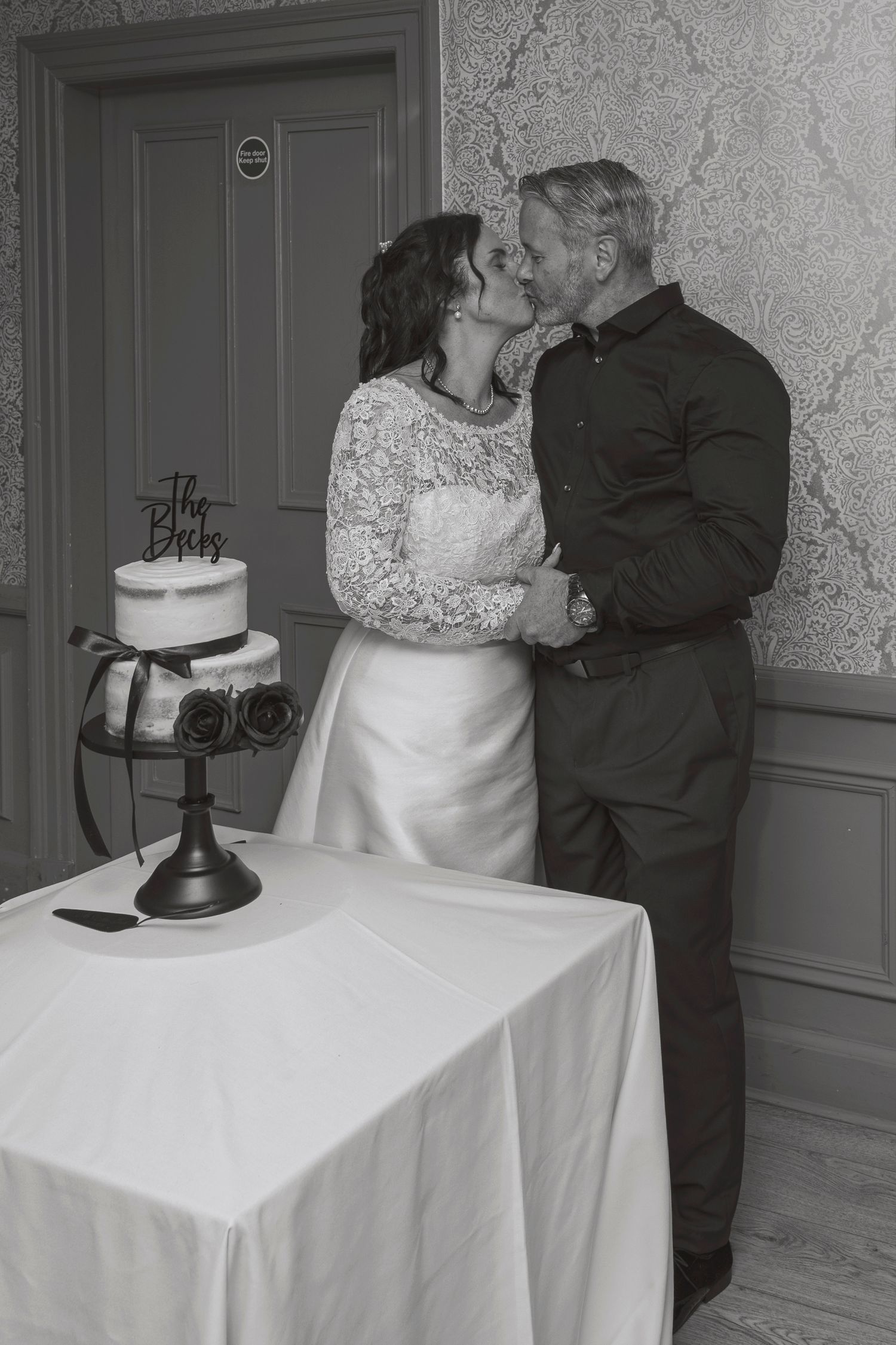 Black and white photo of a bride and groom kissing beside their wedding cake at their reception.