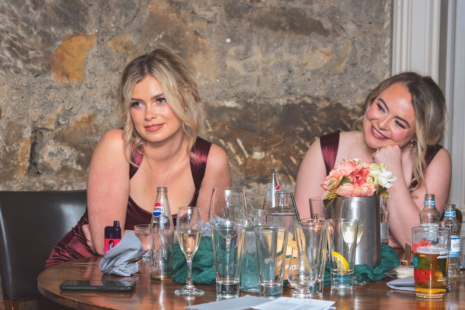Two women in formal attire sitting at a decorated dinner table with glasses and flowers.
