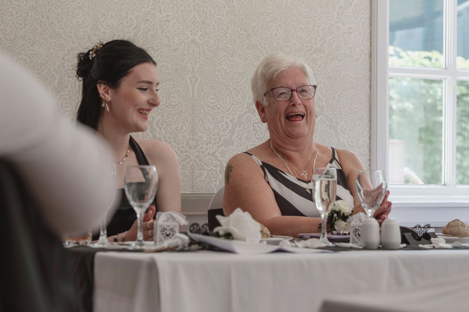 Two women laughing joyfully at a formal dinner table, sharing a warm moment at an elegant event.