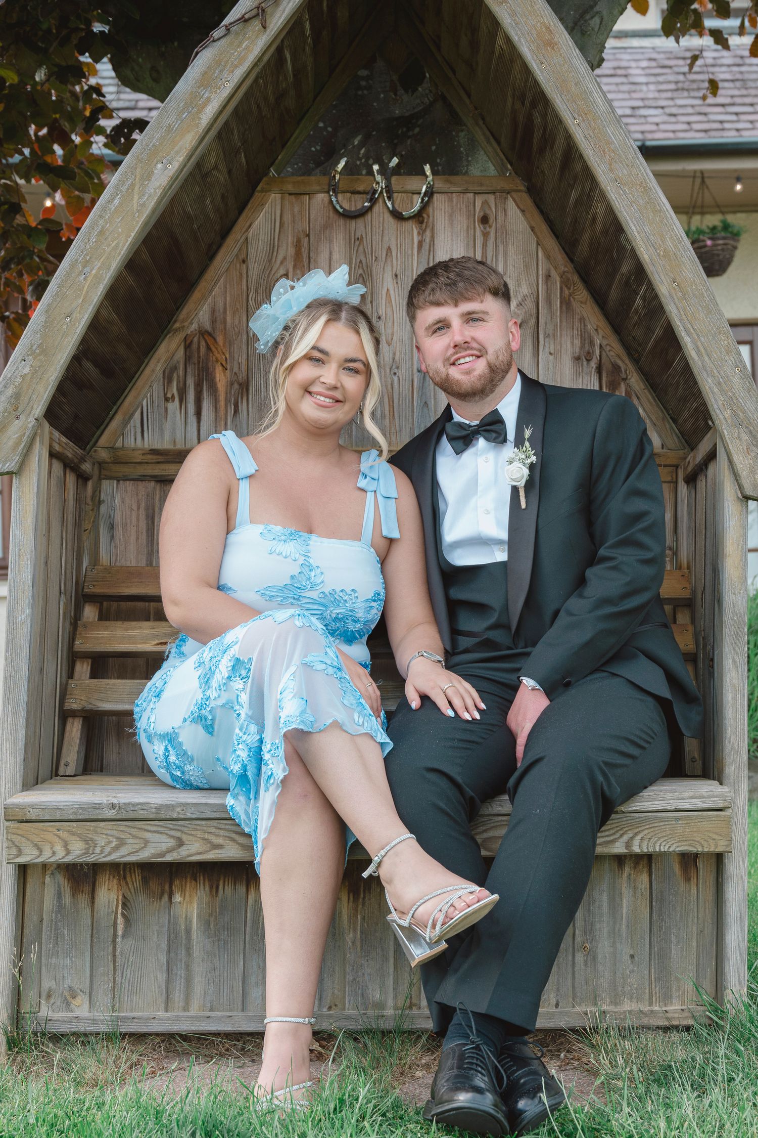 Couple posing in wooden arch gazebo, woman in blue floral dress with hair accessory, man in black tuxedo with boutonniere.