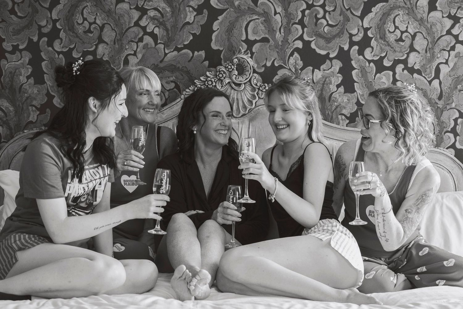Four women sitting on a bed laughing and toasting with champagne glasses in a black and white photo.