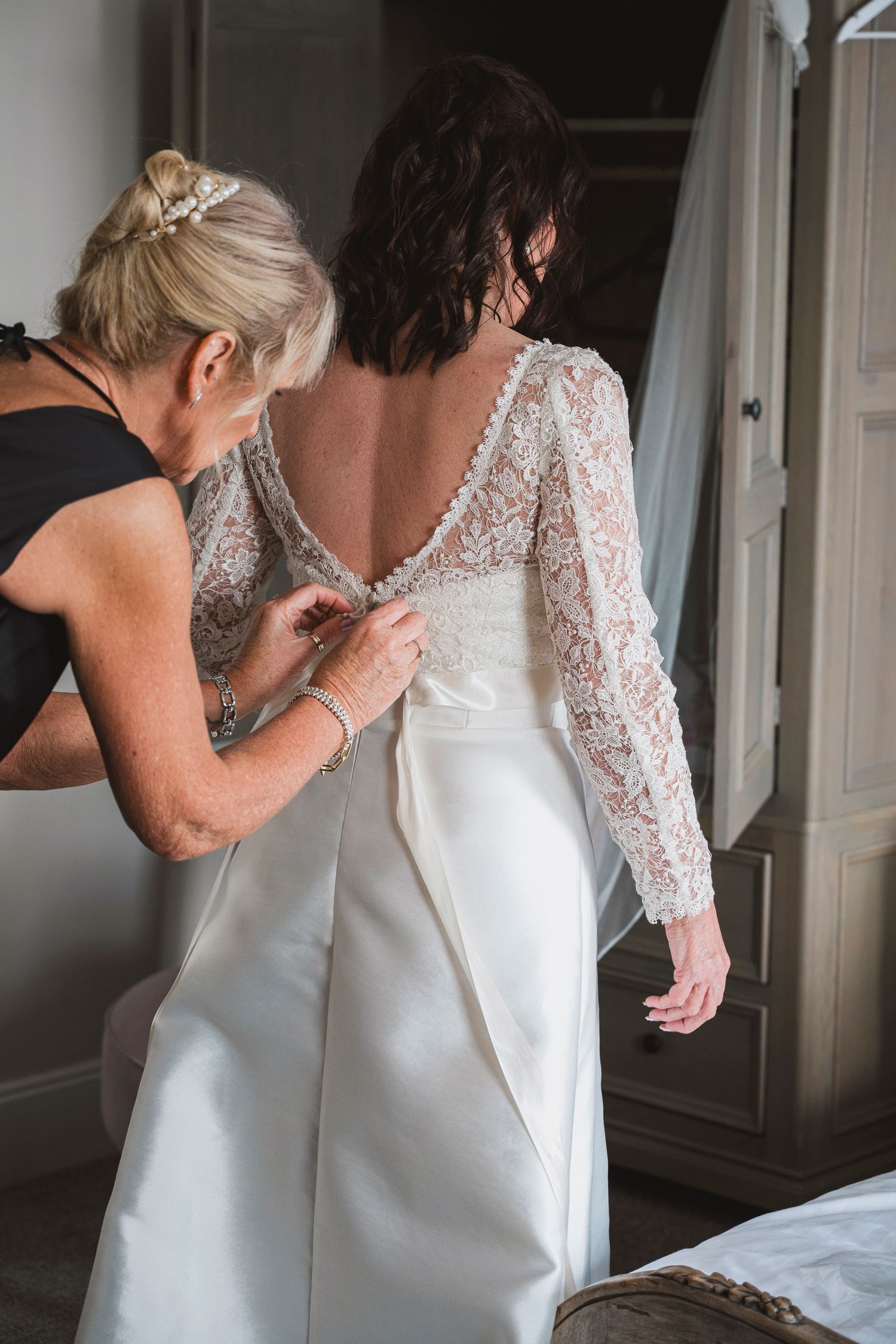 A woman helps button the back of a bride's elegant lace and satin wedding dress before the ceremony.