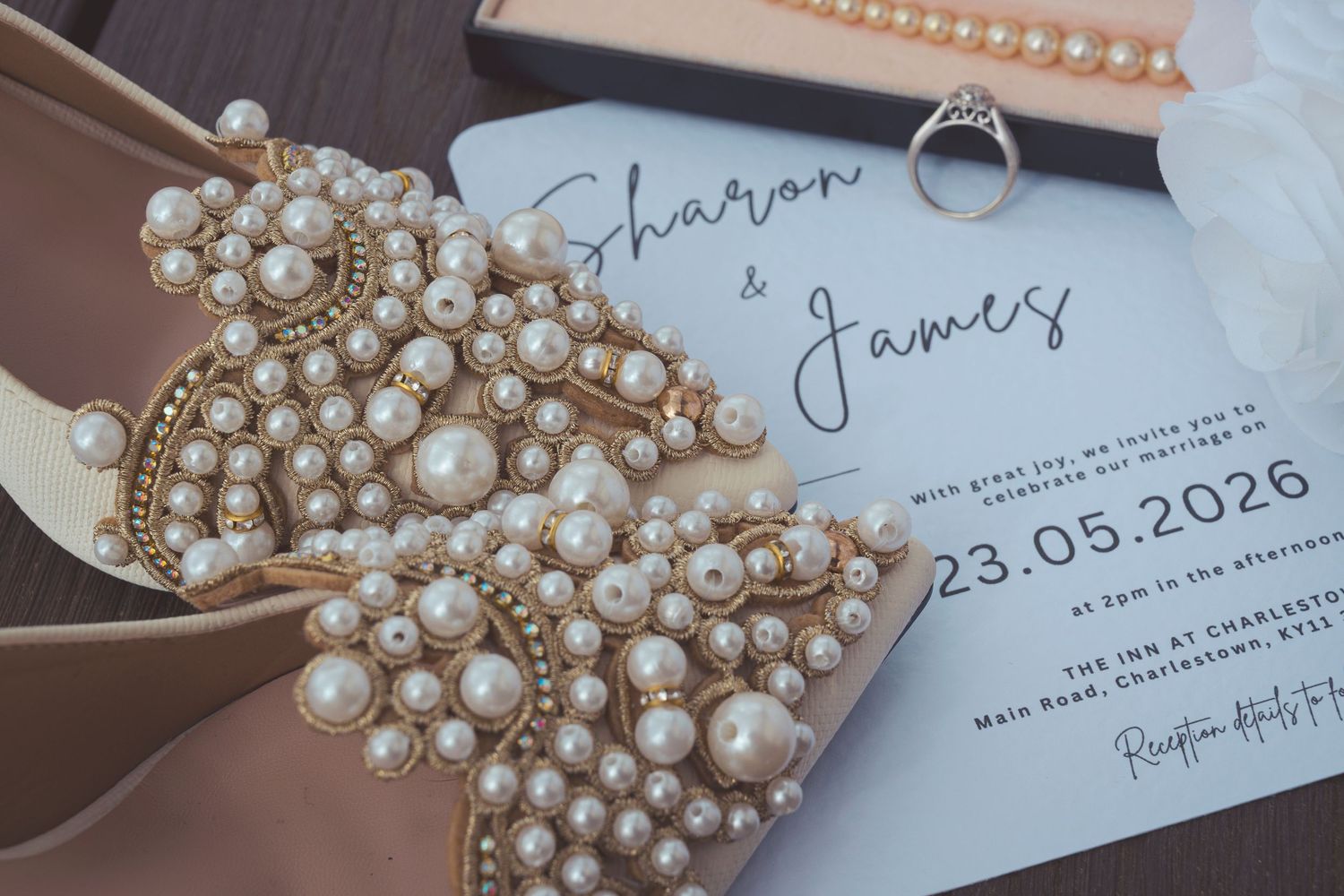 Pearl-embellished bridal shoes beside a wedding invitation for Sharon and James dated 23.05.2026, with a diamond ring and pearl necklace.