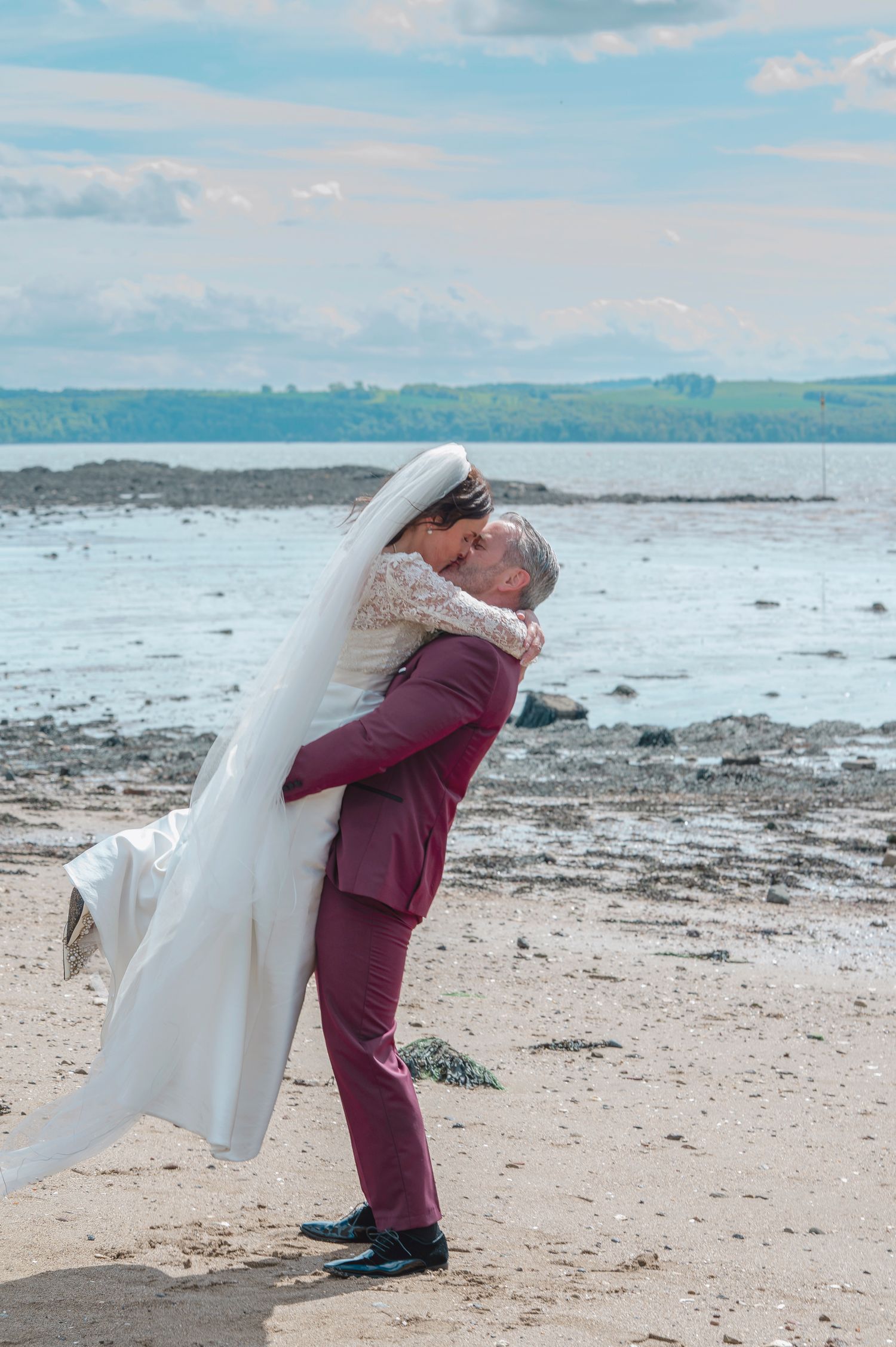 Groom in burgundy suit lifts bride in white gown for a kiss on a sandy beach with ocean views.