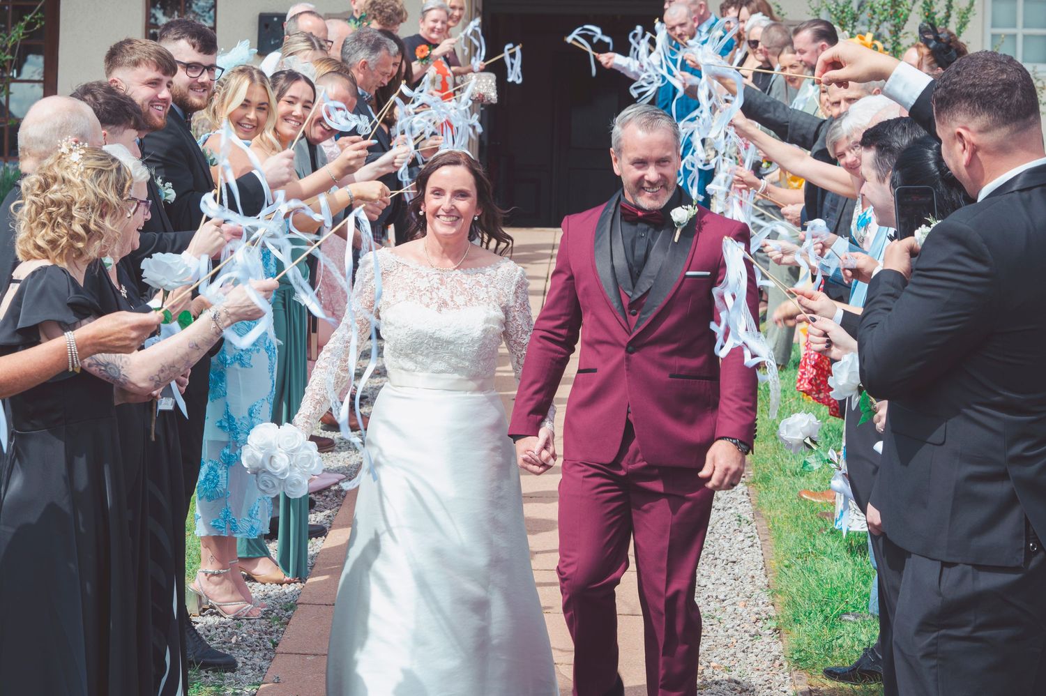 Happy newlyweds walk through a celebratory ribbon tunnel surrounded by smiling wedding guests outdoors.