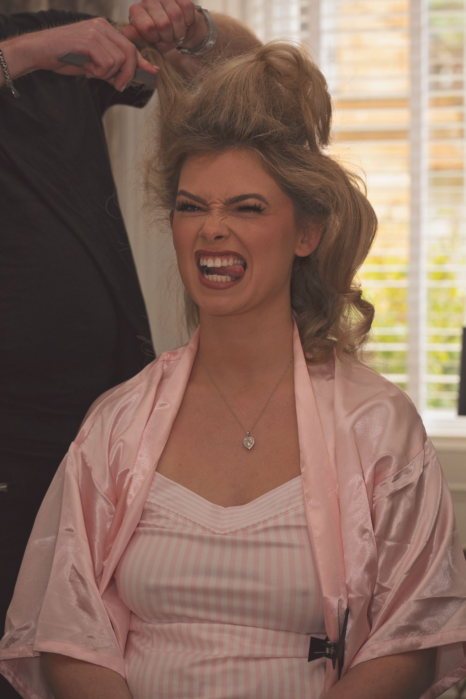 Bride grimacing in pain while getting her blonde hair styled in an elegant updo, wearing a pink satin robe.