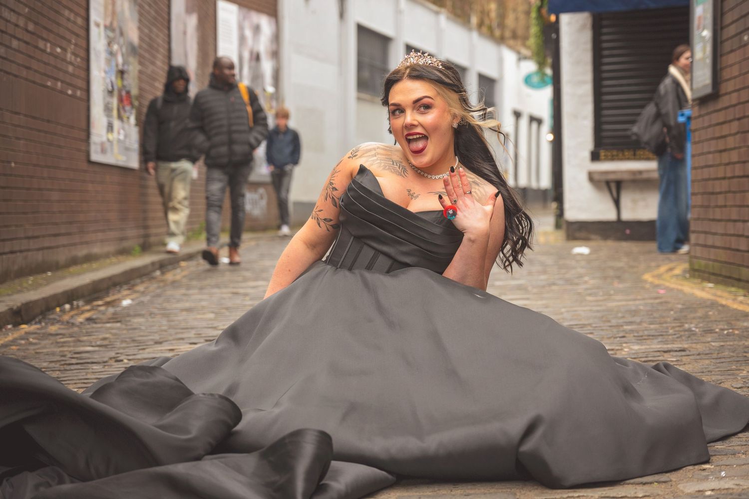 A joyful bride in a dramatic black ball gown sits on a cobblestone street laughing outdoors.