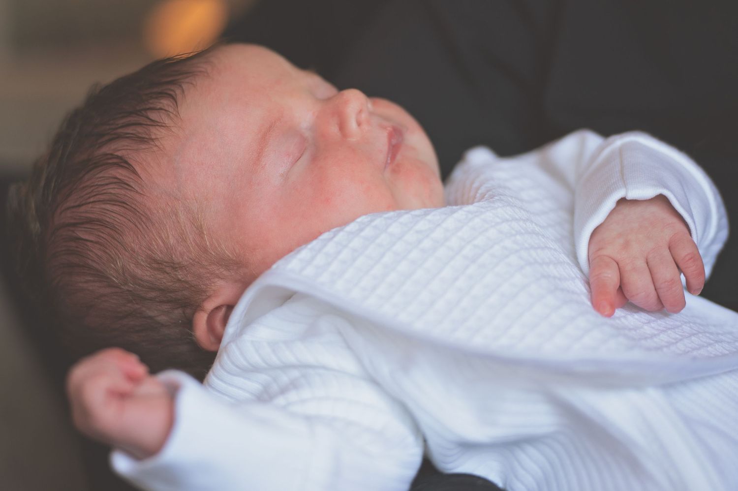 Sleeping newborn baby wrapped in white blanket lying on soft surface in natural light.
