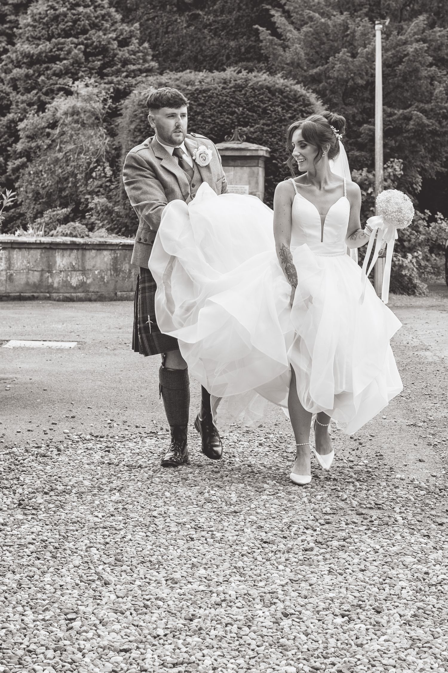 Black and white photo of couple in wedding attire with dress flowing in wind on gravel path.