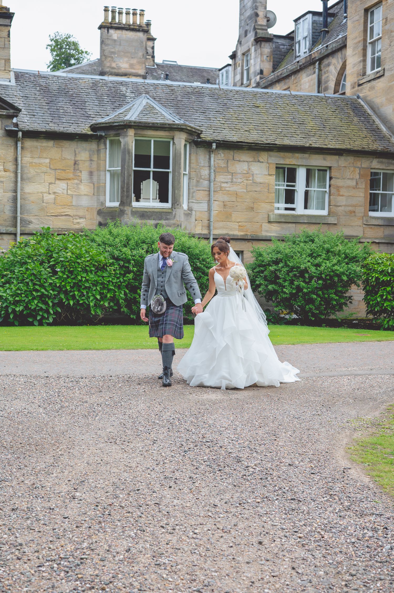 Wedding couple walking together on gravel path with traditional Scottish attire and stone building backdrop.