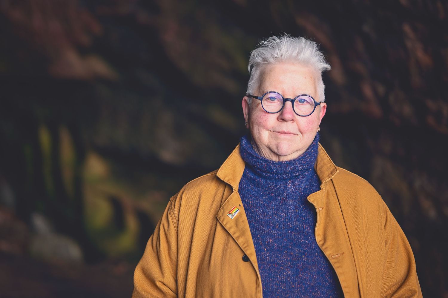 The Author Val Mcdermid with short white hair and round glasses wearing a mustard jacket and blue turtleneck sweater in the Weymss caves