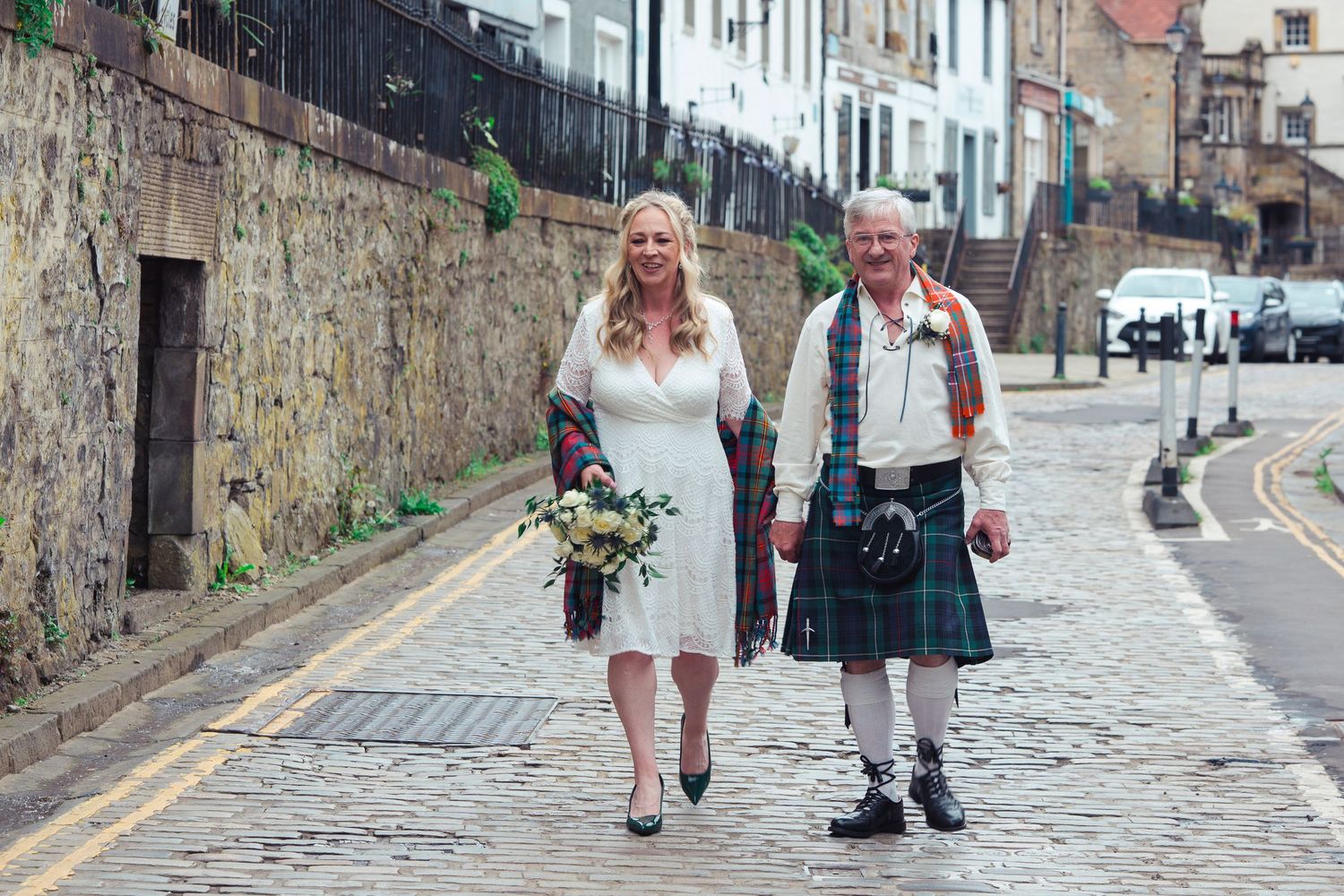 Bride in white dress and groom in traditional Scottish kilt walking together on cobblestone street on their wedding day.
