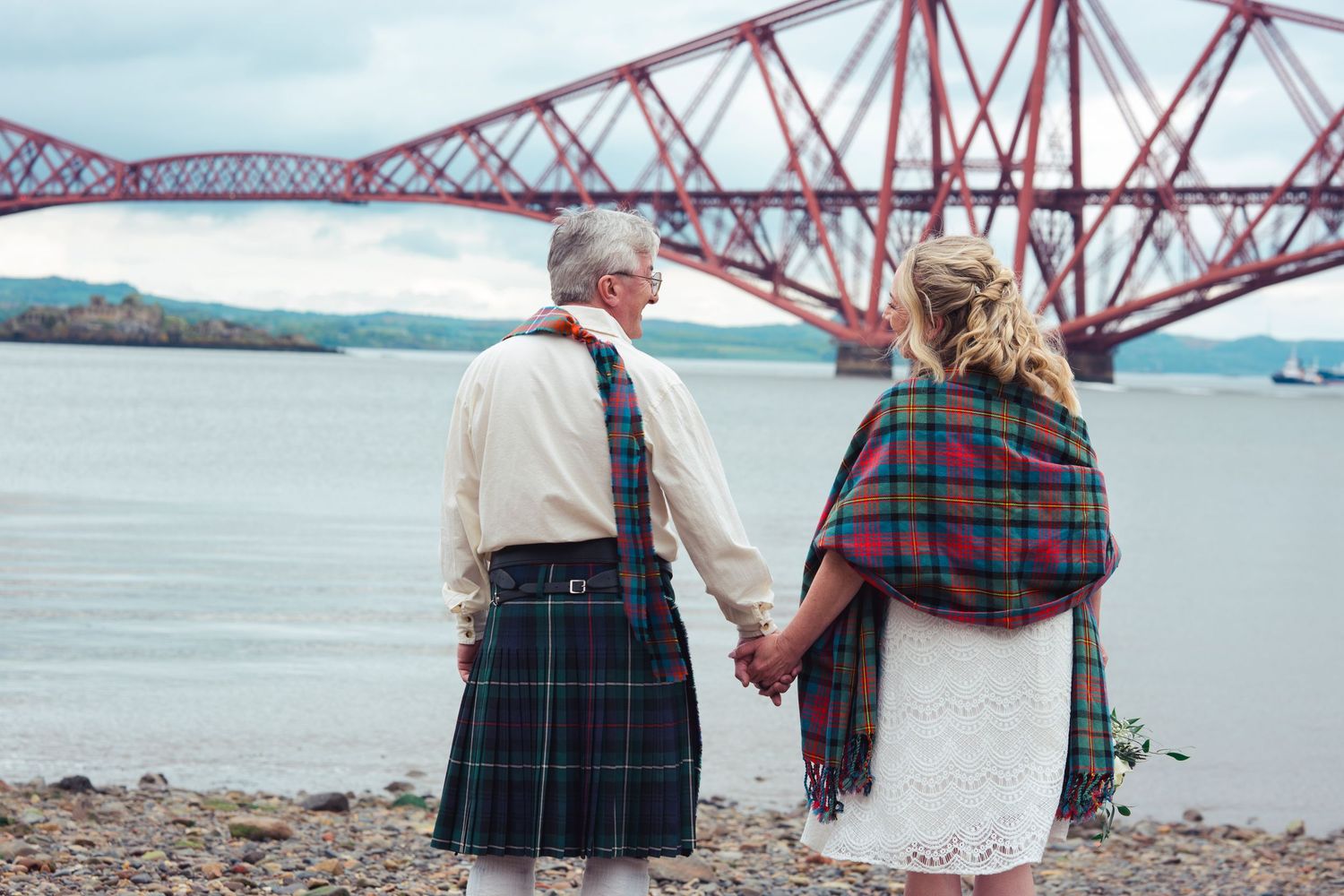 Couple in Scottish tartan holding hands by the Forth Bridge, Edinburgh, on their wedding day.