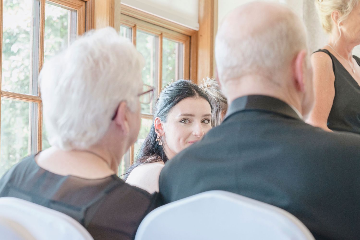 A dark-haired woman smiles warmly at an elderly couple during a wedding ceremony indoors.