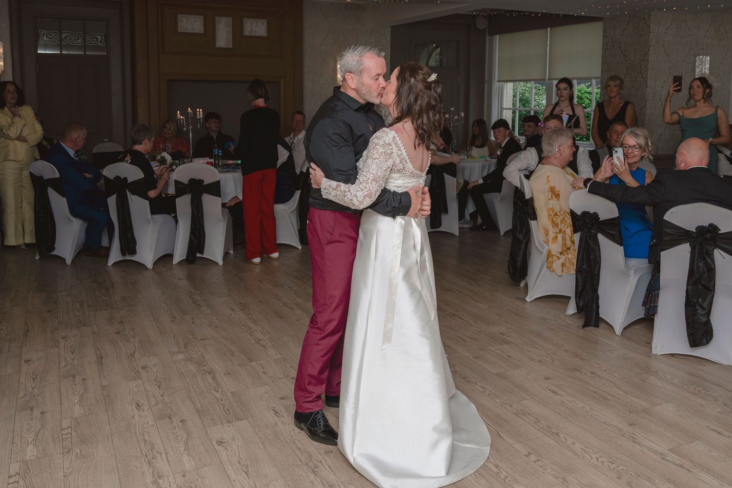 Bride and groom share first dance kiss at wedding reception with guests seated at decorated tables.