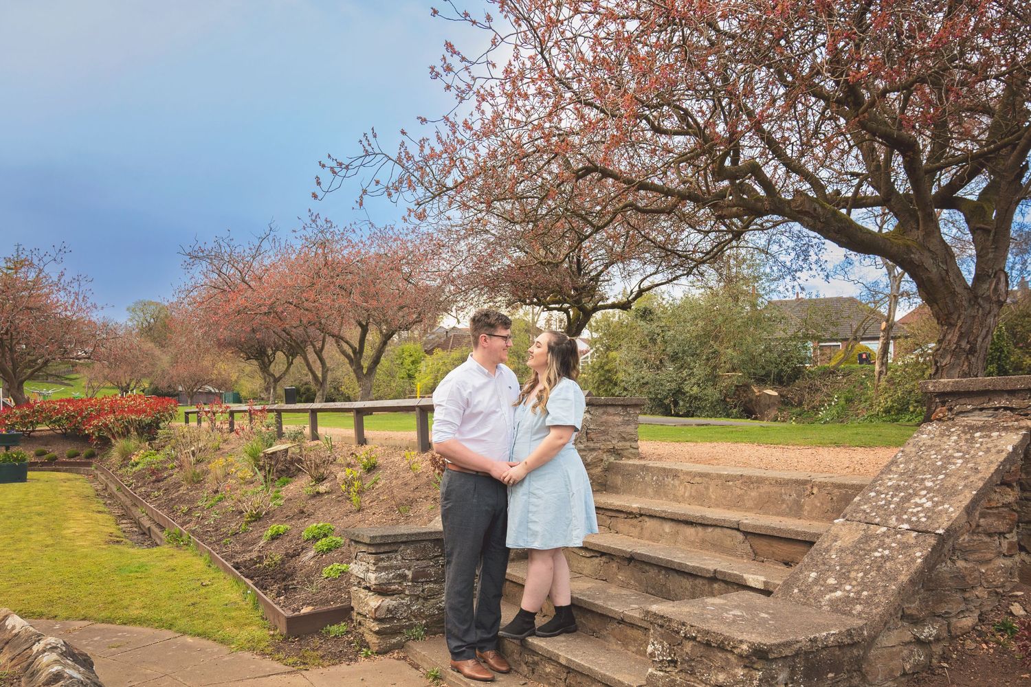 Couple standing together in a garden with cherry blossom trees and stone steps under a dramatic blue sky.