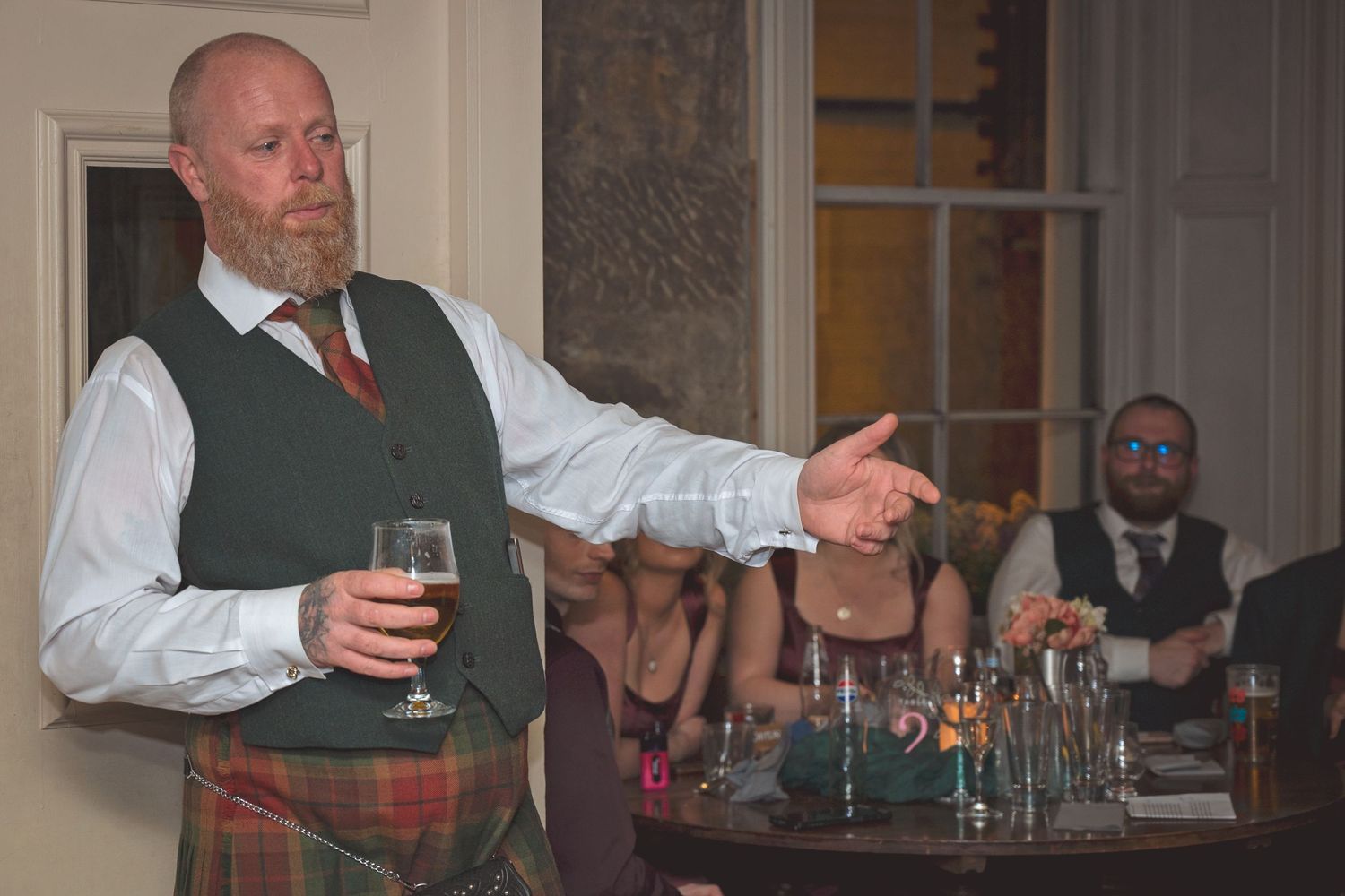 Scottish man in tartan kilt and green waistcoat giving speech holding pint glass at wedding.