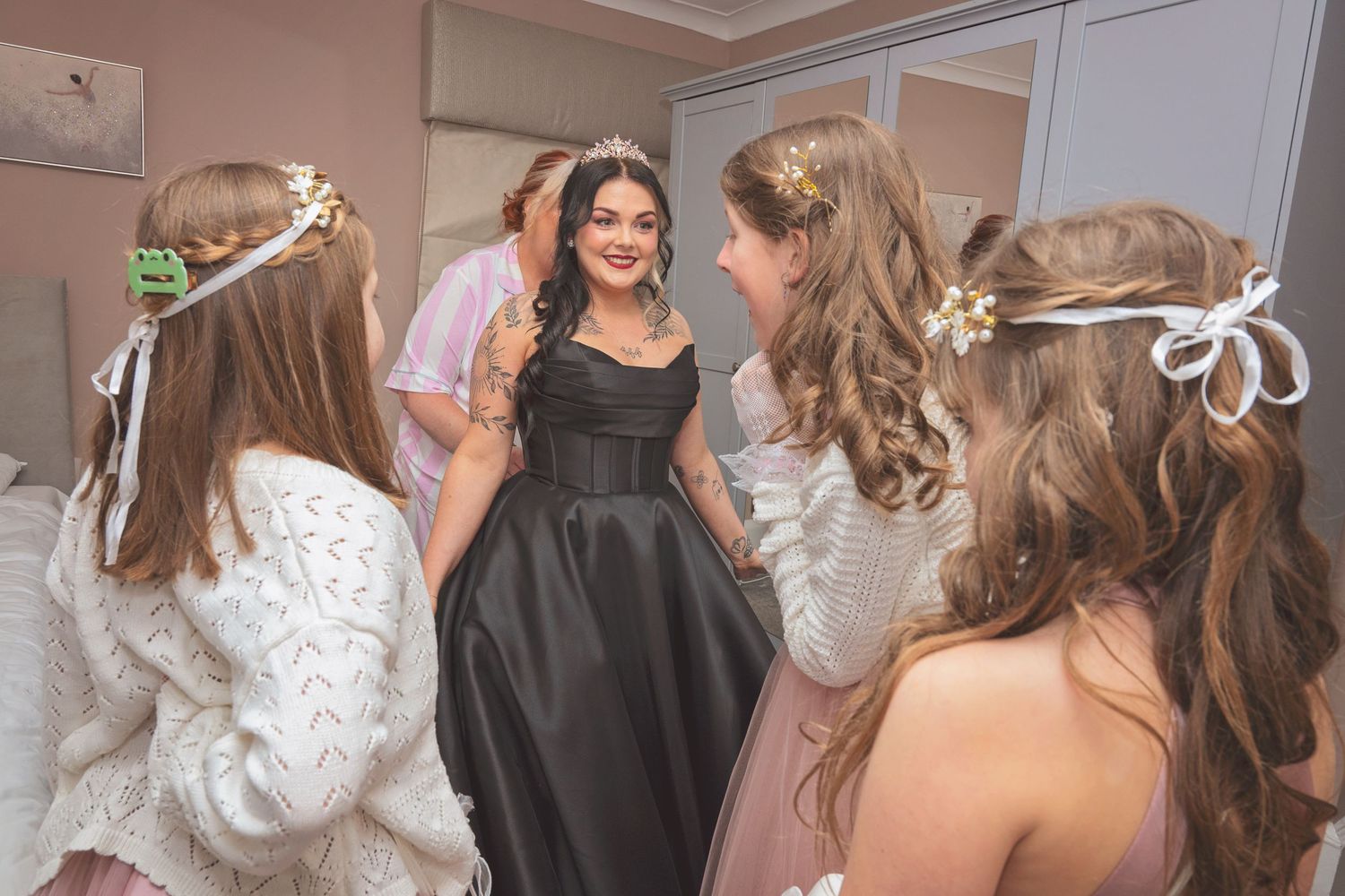 A smiling young woman in a black gown and tiara is surrounded by bridesmaids wearing floral hair accessories.