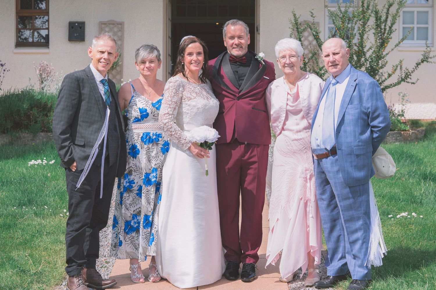 A bride and groom pose with four family members outside a white building on a sunny wedding day.