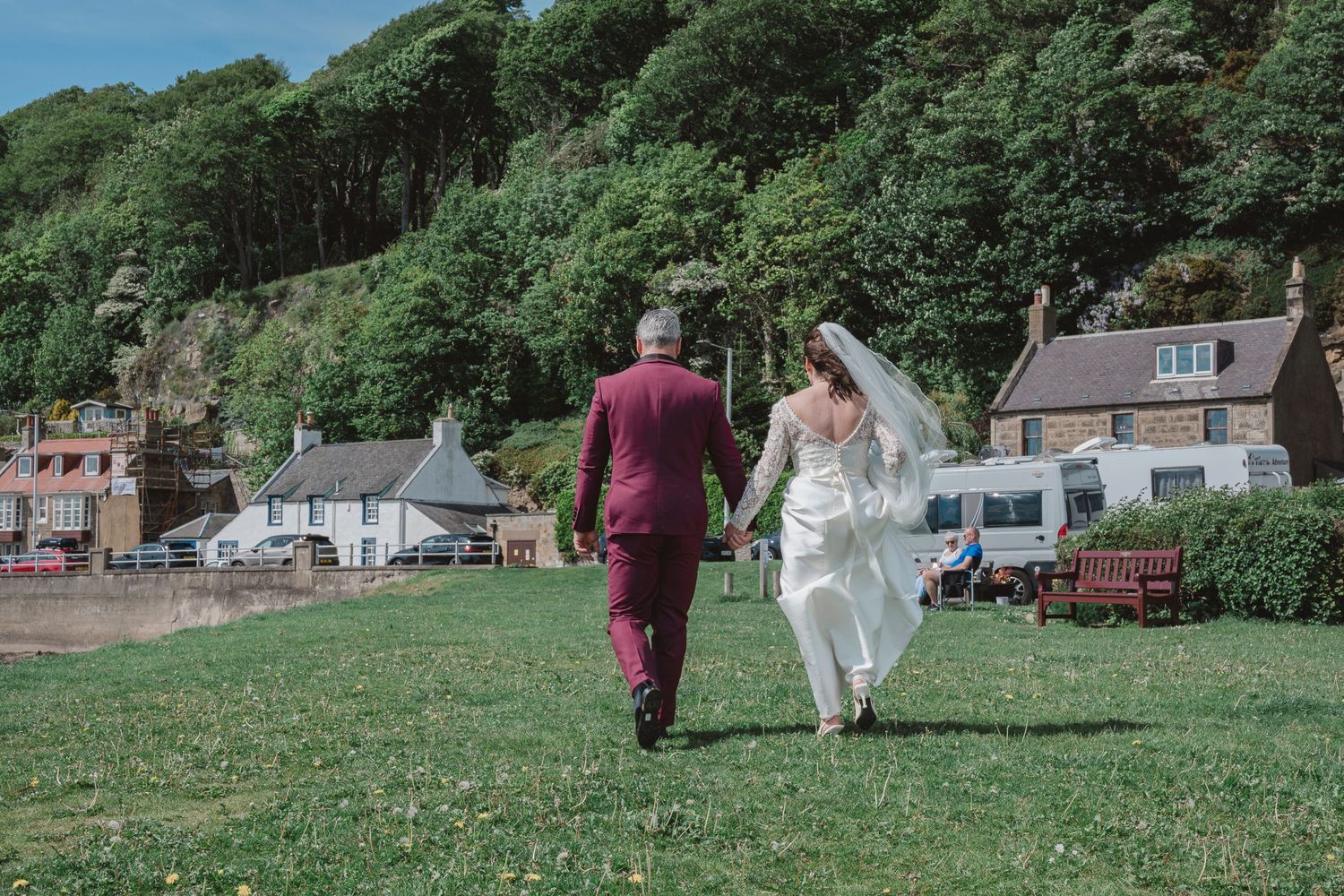 A bride and groom walk hand in hand across green grass toward a quaint village with trees and cottages in the background.