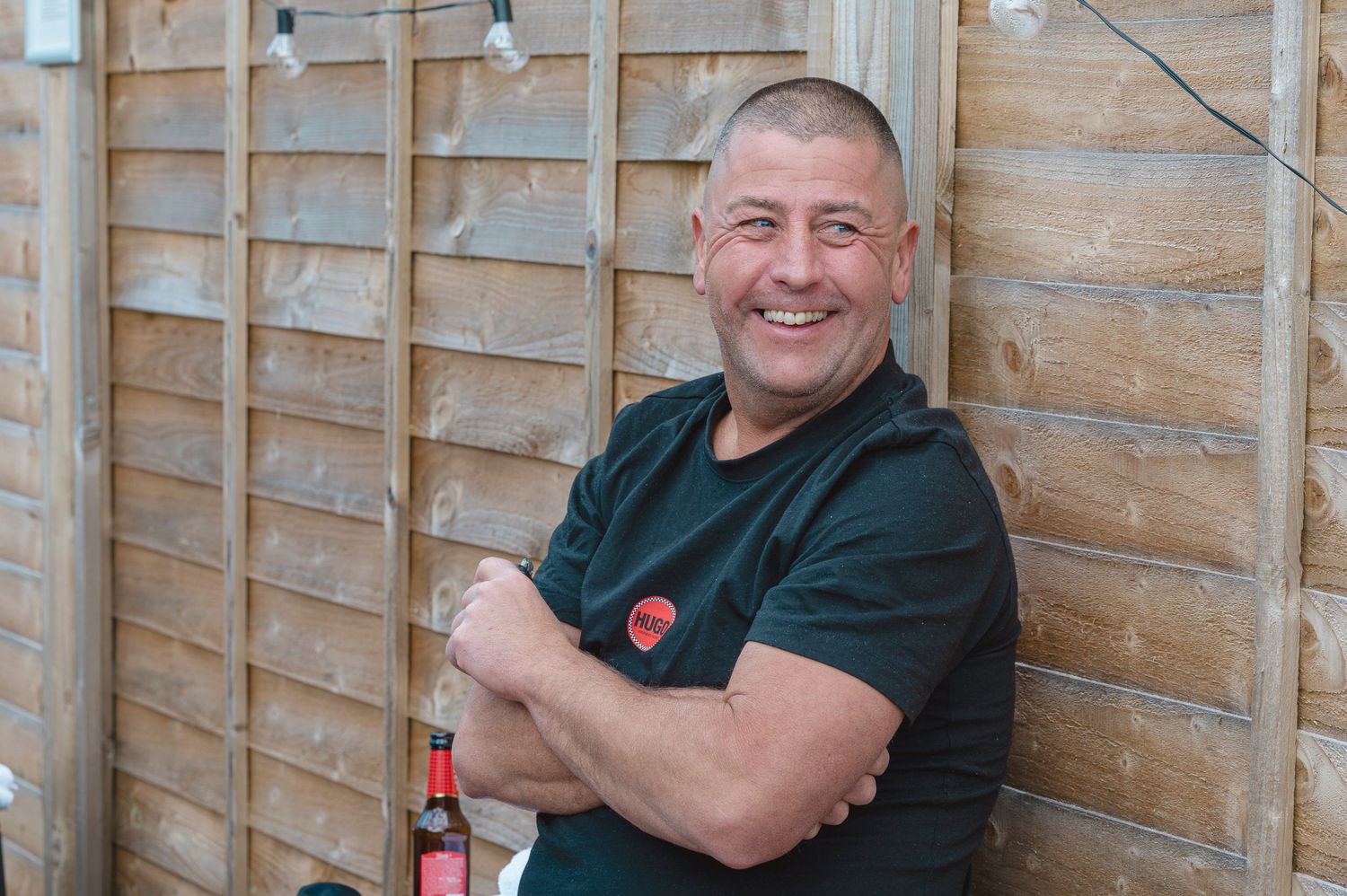 Smiling man with crossed arms holding a beer bottle, leaning against a wooden fence outdoors.